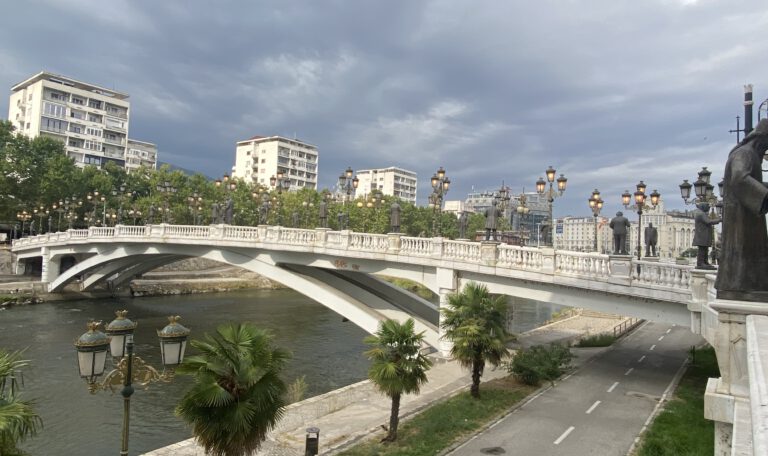 На фотографии изображён один из самых известных мостов Скопье — Каменный мост, перекинутый через реку Вардар. Вдоль моста установлены высокие изящные фонари в классическом стиле, которые вечером красиво подсвечиваются тёплым светом. По бокам стоят массивные статуи в античном духе, придающие мосту монументальность. На заднем плане виднеются здания, выполненные в стиле "неоклассика", и голубое небо, отражающееся в воде. Атмосфера на фото — торжественная и спокойная, передающая дух города, где история встречается с современностью.