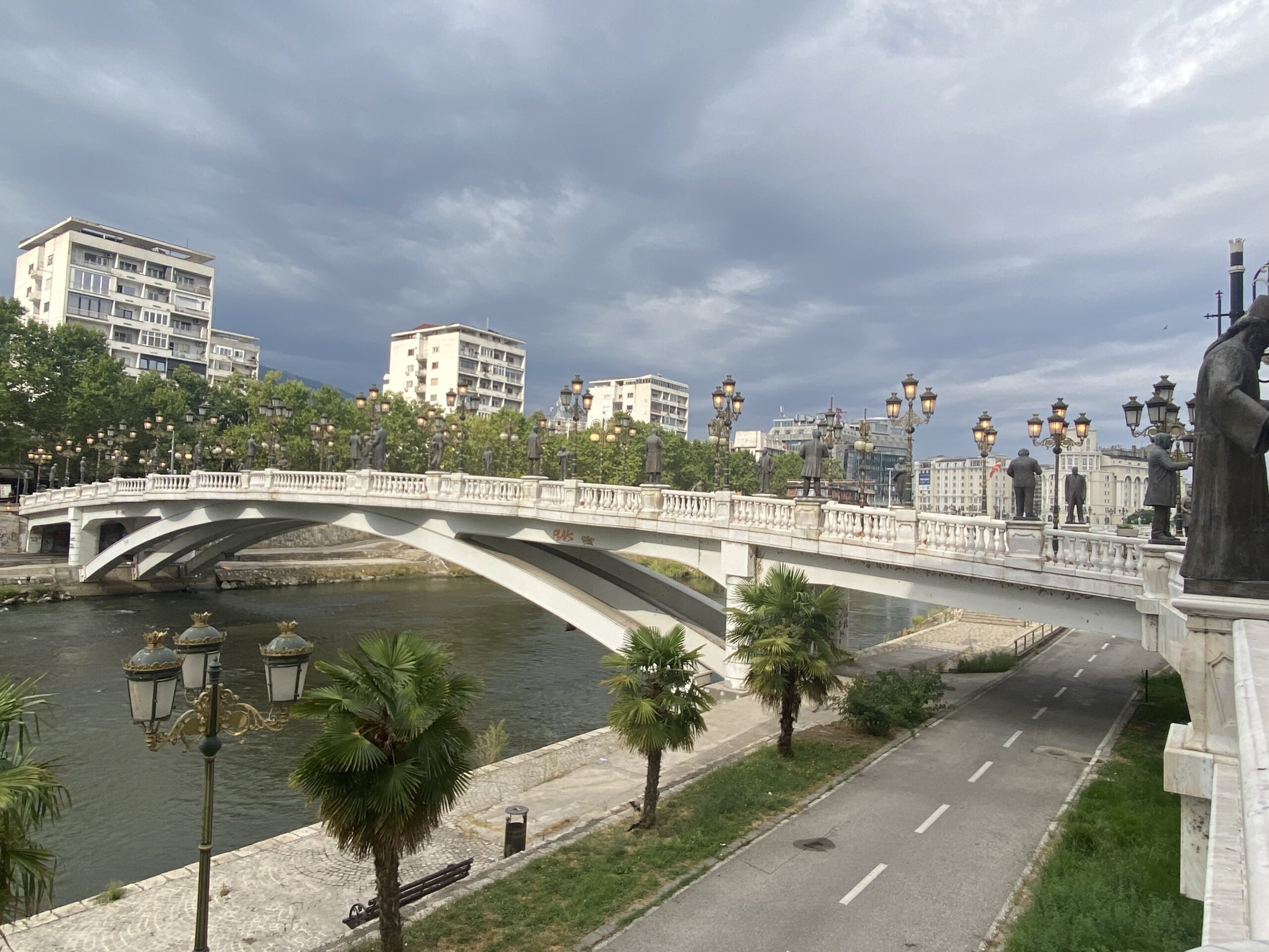 На фотографии изображён один из самых известных мостов Скопье — Каменный мост, перекинутый через реку Вардар. Вдоль моста установлены высокие изящные фонари в классическом стиле, которые вечером красиво подсвечиваются тёплым светом. По бокам стоят массивные статуи в античном духе, придающие мосту монументальность. На заднем плане виднеются здания, выполненные в стиле "неоклассика", и голубое небо, отражающееся в воде. Атмосфера на фото — торжественная и спокойная, передающая дух города, где история встречается с современностью.