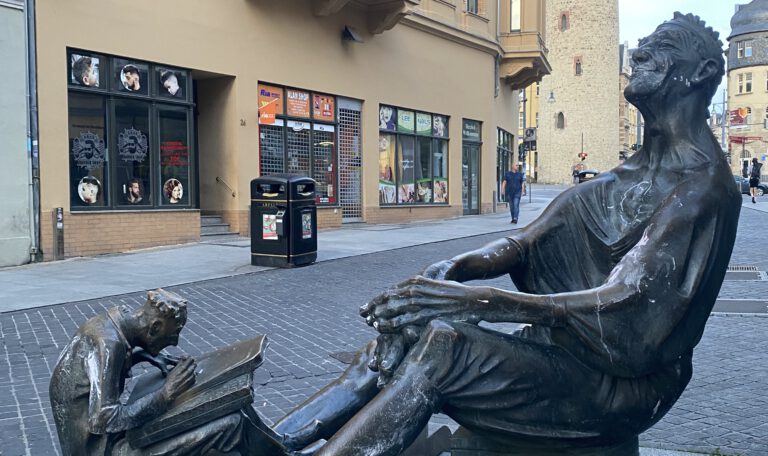 Бронзовая скульптура на тихой улице в Халле: взрослый мужчина сидит, спокойно наблюдая, а у его ног мальчик сосредоточенно читает книгу. Атмосфера тишины и размышления.