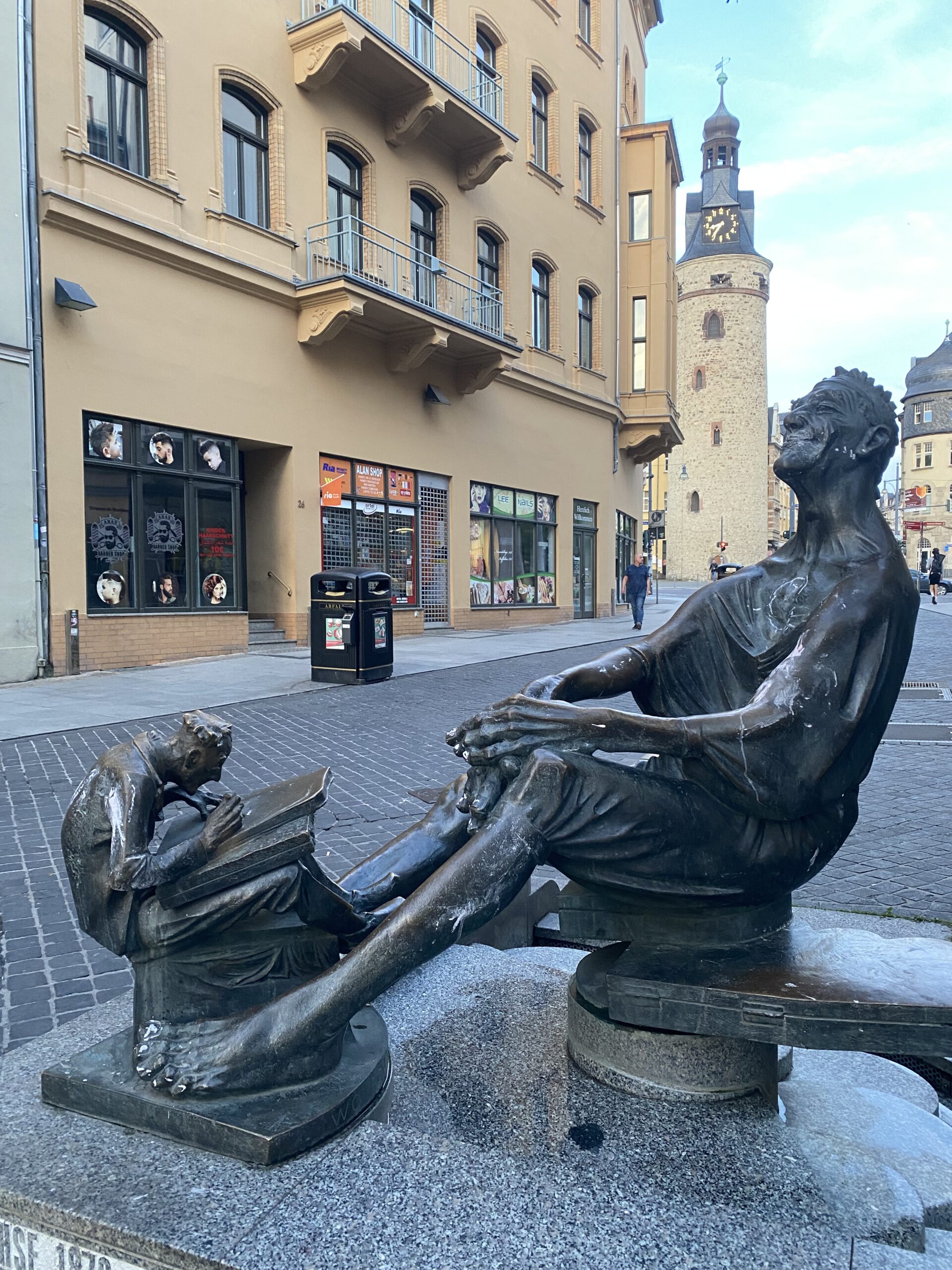 Бронзовая скульптура на тихой улице в Халле: взрослый мужчина сидит, спокойно наблюдая, а у его ног мальчик сосредоточенно читает книгу. Атмосфера тишины и размышления.