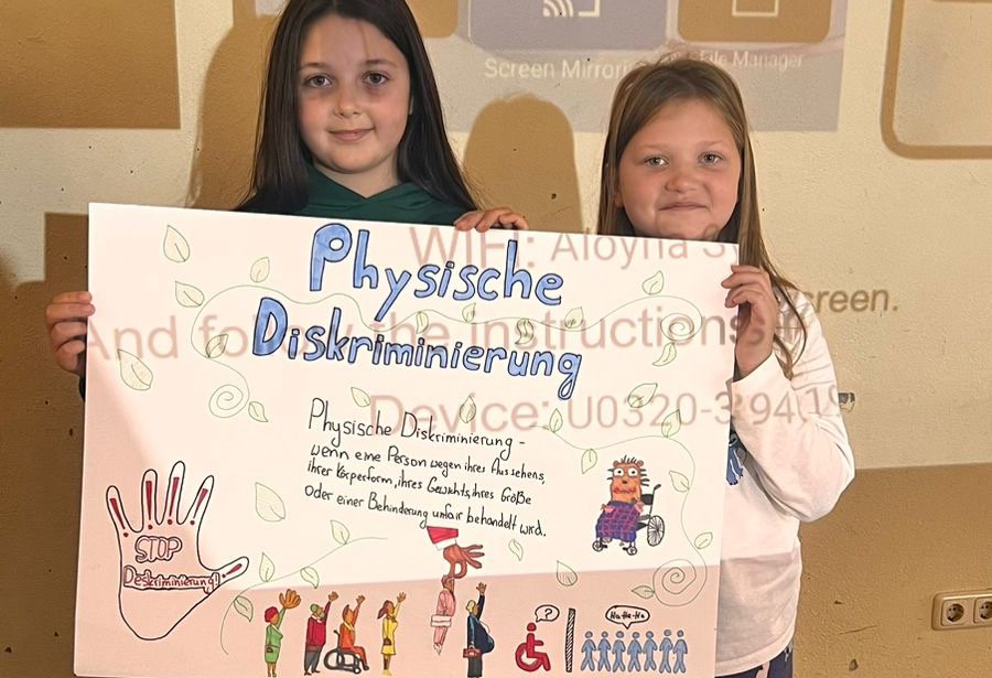zwei Schülerinnen halten eine A2-Papier-Seite mit selbstgemalten Bildern und selbstgeschriebenem Text in den Händen für ihre Präsentation zum Thema Physische Diskriminierung