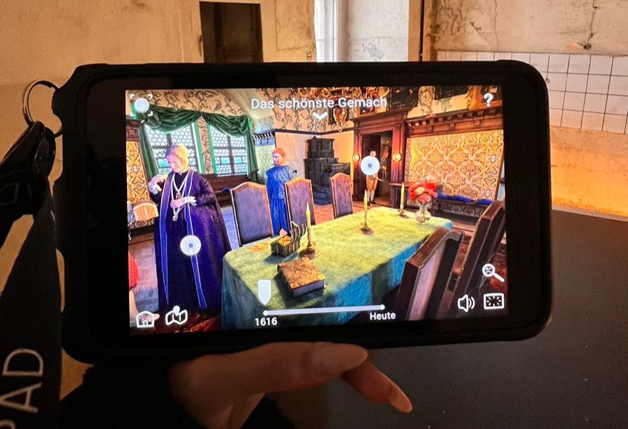 eine Teilnehmerin hält während des interaktiven Rundgangs im Schloss Colditz ein HistoPad in der Hand, auf welchem ein fiktives Foto zu sehen ist, das die ehemalige adlige Bewohner:innen in einem prunkvollen Raum zeigt