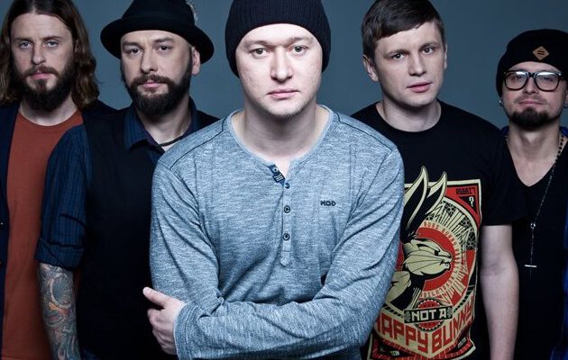 Групповой портрет пяти мужчин, участников группы «Бумбокс», на сером фоне. В центре — солист Андрей Хлывнюк в сером лонгсливе и черной шапке, со сложенными на груди руками. Слева от него стоят двое мужчин: один с татуировками на руках, другой в черной шляпе и жилетке. Справа — двое мужчин в черных футболках, один из которых в очках и шапке. У всех участников спокойные и серьезные выражения лиц.