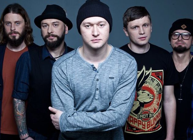 Групповой портрет пяти мужчин, участников группы «Бумбокс», на сером фоне. В центре — солист Андрей Хлывнюк в сером лонгсливе и черной шапке, со сложенными на груди руками. Слева от него стоят двое мужчин: один с татуировками на руках, другой в черной шляпе и жилетке. Справа — двое мужчин в черных футболках, один из которых в очках и шапке. У всех участников спокойные и серьезные выражения лиц.