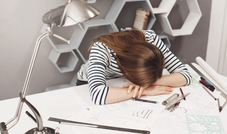 Девушка сидит за столом и, устав, положила голову на него. На столе стоит лампа. Рядом лежат тетради, листочки и ручки.