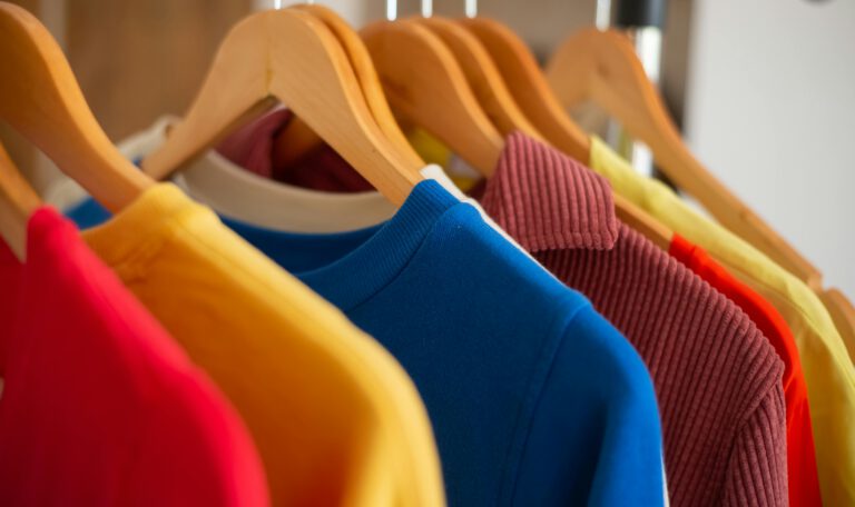 На фотографии запечатлен крупный план яркой одежды, висящей на деревянных плечиках. Снимок сделан под углом, создавая ритмичный ряд уходящих вглубь вешалок. Центральный объект: В фокусе находится ярко-синий хлопковый джемпер с классическим круглым вырезом в рубчик. Его текстура мягкая и плотная. Цветовая палитра: Слева от синего джемпера виден край насыщенно-желтой кофты и ярко-красной ткани. Справа — джемпер глубокого бордового (винного) цвета с вельветовой или трикотажной текстурой в вертикальную полоску, а за ним следуют вещи оранжевого и светло-желтого оттенков. Детали: Вешалки выполнены из светлого гладкого дерева. Они висят на темной металлической перекладине, которая видна в верхней правой части кадра. Освещение и глубина: Освещение мягкое и естественное, падает слева. Передний план и объекты вдали слегка размыты, что заставляет взгляд сфокусироваться на сочном синем цвете и фактуре ткани в центре кадра.