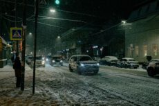 На фотографии запечатлена городская улица в темное время суток во время сильного снегопада. Дорога и тротуары полностью покрыты слоем снега. На переднем плане справа виден современный внедорожник темного цвета с включенными фарами, который едет по заснеженной проезжей части. Вдоль обочин припаркованы автомобили, также засыпанные снегом. Слева на тротуаре, спиной к зрителю, стоит человек в темной зимней одежде. Рядом с ним виден столб с дорожным знаком «Пешеходный переход» на желтом фоне. Улицу освещают фонари, их свет создает ореолы вокруг падающих снежинок. На заднем плане видны силуэты зданий с подсвеченными витринами и огни других машин в потоке. Общая атмосфера снимка передает холод и характерную суету зимнего города в непогоду.