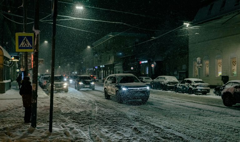 На фотографии запечатлена городская улица в темное время суток во время сильного снегопада. Дорога и тротуары полностью покрыты слоем снега. На переднем плане справа виден современный внедорожник темного цвета с включенными фарами, который едет по заснеженной проезжей части. Вдоль обочин припаркованы автомобили, также засыпанные снегом. Слева на тротуаре, спиной к зрителю, стоит человек в темной зимней одежде. Рядом с ним виден столб с дорожным знаком «Пешеходный переход» на желтом фоне. Улицу освещают фонари, их свет создает ореолы вокруг падающих снежинок. На заднем плане видны силуэты зданий с подсвеченными витринами и огни других машин в потоке. Общая атмосфера снимка передает холод и характерную суету зимнего города в непогоду.