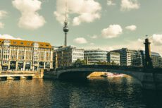 На фотографии запечатлен вид на реку Шпрее в центре Берлина в солнечный день. Основные элементы композиции: Архитектура: На левом берегу реки стоит величественное классическое здание с рыжей черепичной крышей, множеством окон и арочными проемами на первом этаже. Мост: Поперек реки проходит массивный каменный мост с пологими арками, соединяющий берега. Река: На переднем плане — темная вода с легкой рябью, в которой отражаются здания. Вдали под мостом виден небольшой прогулочный катер. Телебашня: На заднем плане, прямо над городскими постройками, возвышается знаменитая Берлинская телебашня с тонким шпилем и шаром наверху. Небо: Светлое, с мягкими белыми облаками, что создает спокойную атмосферу. Изображение выполнено в теплых, слегка приглушенных тонах (эффект ретро-фильтра), что придает городскому пейзажу уютный и исторический вид.