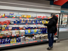 В правой части кадра стоит мужчина среднего телосложения, повернутый спиной и боком к камере. Он одет в теплую черную зимнюю куртку с капюшоном, синие джинсы и темные кроссовки с тремя белыми полосками. Через плечо у него висит черная сумка, а в правой руке он держит синюю пластиковую корзину для покупок. Он внимательно рассматривает товары на верхних полках холодильного стеллажа. Окружение и детали Стеллажи: Перед Денисом расположены длинные открытые полки-холодильники, доверху заполненные продуктами. Полки освещены ярким белым светом. Ассортимент: * На средних полках видны упаковки с моцареллой, пиццей-роллами и различными закусками. Ниже представлены молочные продукты: большие упаковки питьевых йогуртов (маркировка XXL), баночки с десертами и пудингами. На самой нижней полке аккуратно расставлены пачки сливочного масла и маргарина. Ценники: Под каждым товаром видны ярко-красные ценники, которые характерны для периодов распродаж или специальных акций (Aktion). Атмосфера: В магазине чисто, современно и очень светло. Справа видны закрытые стеклянные витрины-холодильники с замороженной продукцией.