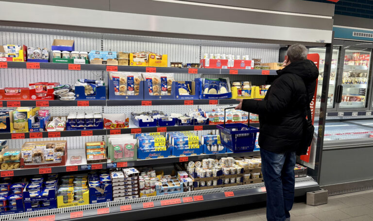 В правой части кадра стоит мужчина среднего телосложения, повернутый спиной и боком к камере. Он одет в теплую черную зимнюю куртку с капюшоном, синие джинсы и темные кроссовки с тремя белыми полосками. Через плечо у него висит черная сумка, а в правой руке он держит синюю пластиковую корзину для покупок. Он внимательно рассматривает товары на верхних полках холодильного стеллажа. Окружение и детали Стеллажи: Перед Денисом расположены длинные открытые полки-холодильники, доверху заполненные продуктами. Полки освещены ярким белым светом. Ассортимент: * На средних полках видны упаковки с моцареллой, пиццей-роллами и различными закусками. Ниже представлены молочные продукты: большие упаковки питьевых йогуртов (маркировка XXL), баночки с десертами и пудингами. На самой нижней полке аккуратно расставлены пачки сливочного масла и маргарина. Ценники: Под каждым товаром видны ярко-красные ценники, которые характерны для периодов распродаж или специальных акций (Aktion). Атмосфера: В магазине чисто, современно и очень светло. Справа видны закрытые стеклянные витрины-холодильники с замороженной продукцией.