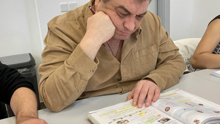 КНа фотографії зображений чоловік зрілого віку, який сидить за столом у світлому навчальному класі. Він зосереджено вивчає розгорнутий підручник. Чоловік одягнений у світлу вельветову сорочку бежевого або пісочного кольору. Однією рукою він підпирає голову, а в іншій тримає червону ручку або маркер, працюючи з текстом. Перед ним на столі лежить велика розгорнута книга з багатьма помітками, зробленими кольоровими маркерами. Поруч розкладене інше навчальне приладдя: різнокольорові фломастери, аркуші з вправами, пенал із квітковим принтом та кабелі для зарядки. Зліва в кадр потрапляє рука іншої людини, яка тримає аркуш паперу, що створює атмосферу групового заняття або мовного курсу. Фон нейтральний: біла стіна, сірі двері та помаранчеве оголошення на них. Загальна атмосфера знімка передає процес глибокого занурення в навчання та концентрацію.