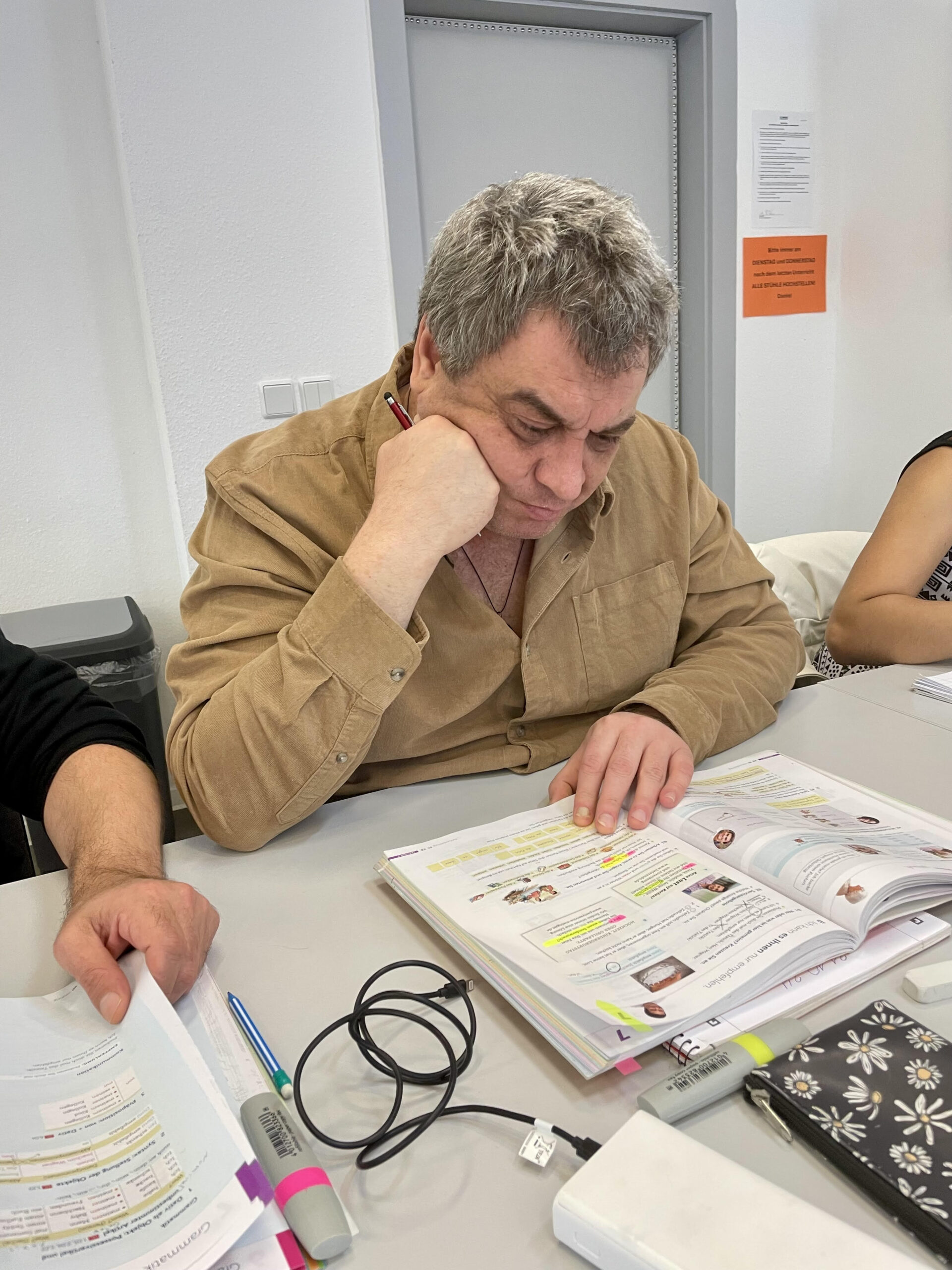 На фотографії зображений чоловік зрілого віку, який сидить за столом у світлому навчальному класі. Він зосереджено вивчає розгорнутий підручник.
Чоловік одягнений у світлу вельветову сорочку бежевого або пісочного кольору. Однією рукою він підпирає голову, а в іншій тримає червону ручку або маркер, працюючи з текстом. Перед ним на столі лежить велика розгорнута книга з багатьма помітками, зробленими кольоровими маркерами. Поруч розкладене інше навчальне приладдя: різнокольорові фломастери, аркуші з вправами, пенал із квітковим принтом та кабелі для зарядки.
Зліва в кадр потрапляє рука іншої людини, яка тримає аркуш паперу, що створює атмосферу групового заняття або мовного курсу. Фон нейтральний: біла стіна, сірі двері та помаранчеве оголошення на них. Загальна атмосфера знімка передає процес глибокого занурення в навчання та концентрацію. КНа фотографії зображений чоловік зрілого віку, який сидить за столом у світлому навчальному класі. Він зосереджено вивчає розгорнутий підручник. Чоловік одягнений у світлу вельветову сорочку бежевого або пісочного кольору. Однією рукою він підпирає голову, а в іншій тримає червону ручку або маркер, працюючи з текстом. Перед ним на столі лежить велика розгорнута книга з багатьма помітками, зробленими кольоровими маркерами. Поруч розкладене інше навчальне приладдя: різнокольорові фломастери, аркуші з вправами, пенал із квітковим принтом та кабелі для зарядки. Зліва в кадр потрапляє рука іншої людини, яка тримає аркуш паперу, що створює атмосферу групового заняття або мовного курсу. Фон нейтральний: біла стіна, сірі двері та помаранчеве оголошення на них. Загальна атмосфера знімка передає процес глибокого занурення в навчання та концентрацію.