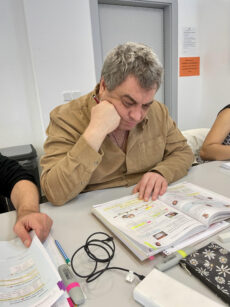 На фотографии запечатлен мужчина зрелого возраста, сидящий за столом в светлом учебном классе. Он сосредоточенно изучает открытый учебник. Мужчина одет в светлую вельветовую рубашку бежевого или песочного цвета. Одной рукой он подпирает голову, а в другой держит красную ручку или маркер, работая с текстом. Перед ним на столе лежит большая раскрытая книга с множеством пометок, сделанных цветными маркерами. Рядом разложены другие учебные принадлежности: разноцветные фломастеры, листы с упражнениями, пенал с цветочным принтом и зарядные кабели. Слева в кадр попадает рука другого человека, который держит лист бумаги, что создает атмосферу группового занятия или языкового курса. Фон нейтральный: белая стена, серая дверь и оранжевое объявление на ней. Общая атмосфера снимка передает процесс глубокого погружения в учебу и концентрацию.