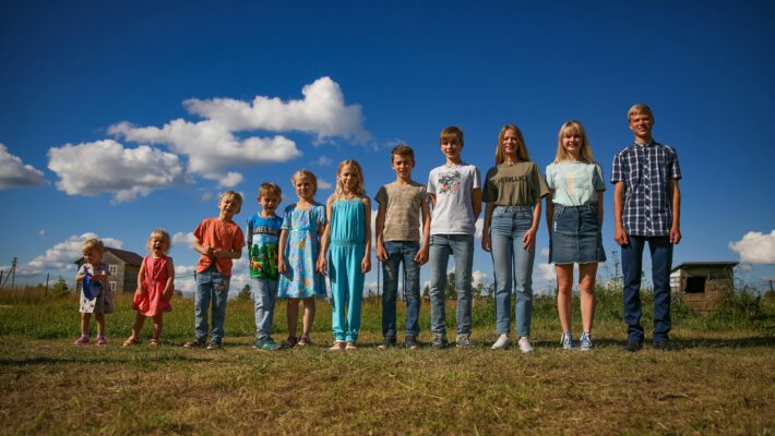 Группа детей и подростков разного возраста, от малышей до старшеклассников, стоят в ряд по росту на фоне открытого поля и голубого неба. Фотография символизирует этапы взросления и смену возрастных категорий