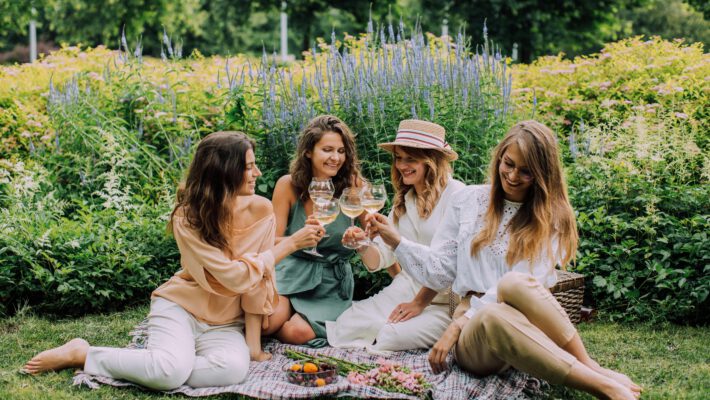 На фотографии запечатлена уютная сцена пикника четырех молодых женщин-подруг в пышном зеленом саду или парке. Они сидят босиком на мягком клетчатом одеяле, расстеленном на траве, и радостно чокаются бокалами с белым вином в тосте. Все девушки одеты в стильную летнюю одежду светлых оттенков (персиковый, шалфейный, белый), а одна из них — в соломенной шляпе-канотье. Позади них раскинулась великолепная клумба с густыми зарослями цветов, включая tall лавандово-синие колосья шалфея и цветущие желтые кустарники. Перед ними на одеяле стоят чаши со свежей клубникой, черешней и лежит букет полевых цветов. Атмосфера фото наполнена смехом, теплом и расслабленным отдыхом в окружении природы.