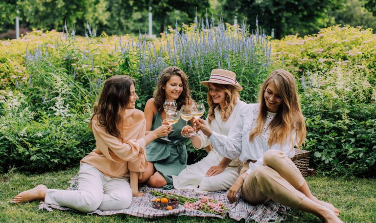 На фотографии запечатлена уютная сцена пикника четырех молодых женщин-подруг в пышном зеленом саду или парке. Они сидят босиком на мягком клетчатом одеяле, расстеленном на траве, и радостно чокаются бокалами с белым вином в тосте. Все девушки одеты в стильную летнюю одежду светлых оттенков (персиковый, шалфейный, белый), а одна из них — в соломенной шляпе-канотье. Позади них раскинулась великолепная клумба с густыми зарослями цветов, включая tall лавандово-синие колосья шалфея и цветущие желтые кустарники. Перед ними на одеяле стоят чаши со свежей клубникой, черешней и лежит букет полевых цветов. Атмосфера фото наполнена смехом, теплом и расслабленным отдыхом в окружении природы.