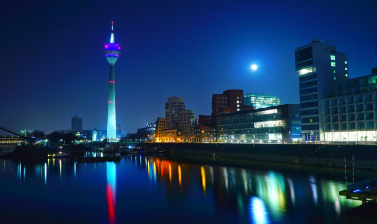 Ночной Дюссельдорф, Германия: подсвеченная телебашня Рейнтурм и современные офисные здания, отражающиеся в реке Рейн. Символ технологического прогресса и цифровизации.