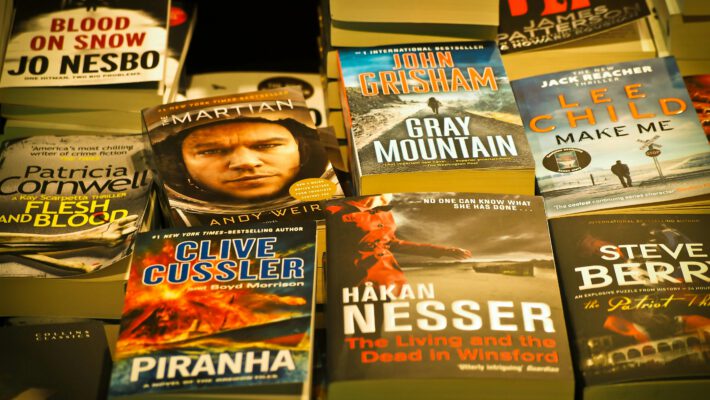 На фотографии запечатлена раскладка популярных современных книг в мягком переплете, лежащих на прилавке или полке. Книги расположены плотными рядами, частично перекрывая друг друга, что создает атмосферу книжного магазина или ярмарки