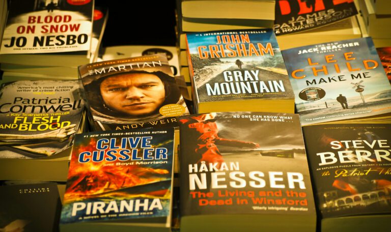 На фотографии запечатлена раскладка популярных современных книг в мягком переплете, лежащих на прилавке или полке. Книги расположены плотными рядами, частично перекрывая друг друга, что создает атмосферу книжного магазина или ярмарки