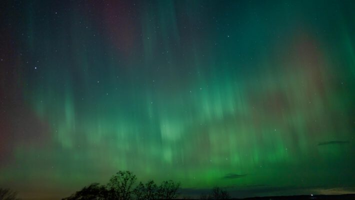 Сильное северное сияние (Aurora Borealis) уровня G3, вызванное геомагнитной бурей, над горизонтом с деревьями и озером. Пример того, как магнитная буря G3 делает полярное сияние видимым в средних широтах Европы.