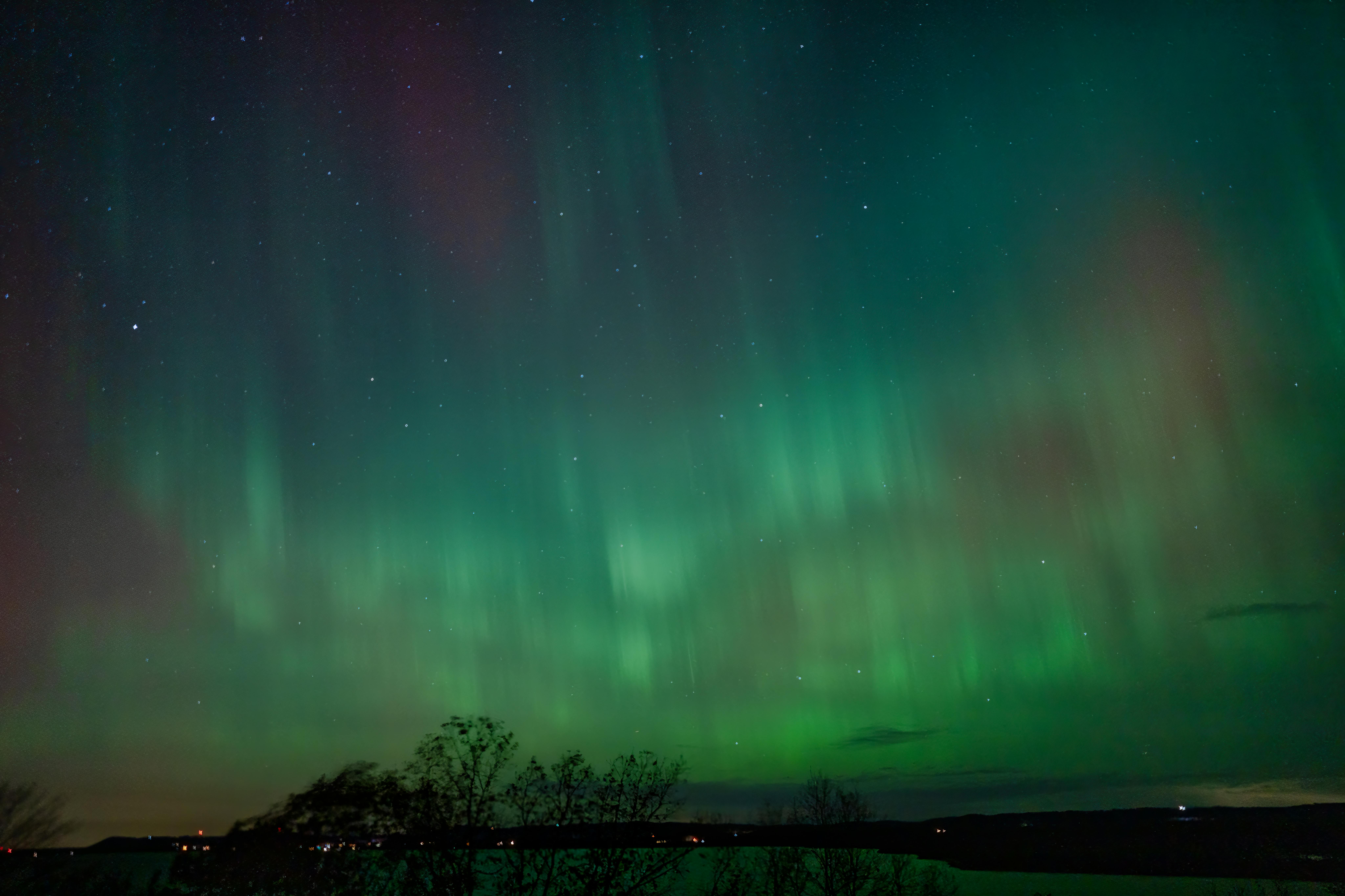 Сильное северное сияние (Aurora Borealis) уровня G3, вызванное геомагнитной бурей, над горизонтом с деревьями и озером. Пример того, как магнитная буря G3 делает полярное сияние видимым в средних широтах Европы.