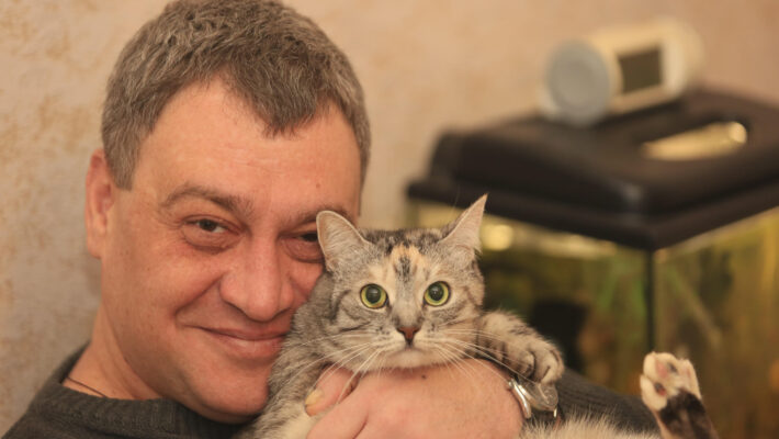 Узнайте, как работает кошачья память и могут ли кошки забывать своих владельцев. Портретный снимок крупным планом, на котором запечатлен радостный мужчина средних лет в сером вязаном свитере, ласково прижимающий к щеке полосатую серую кошку. Кот с любопытством смотрит прямо в объектив, широко раскрыв зеленые глаза, а мужчина тепло улыбается. Рука мужчины с часами на запястье бережно держит питомца, чья лапка видна на переднем плане. На заднем плане размыто виднеются аквариум и другие предметы интерьера
