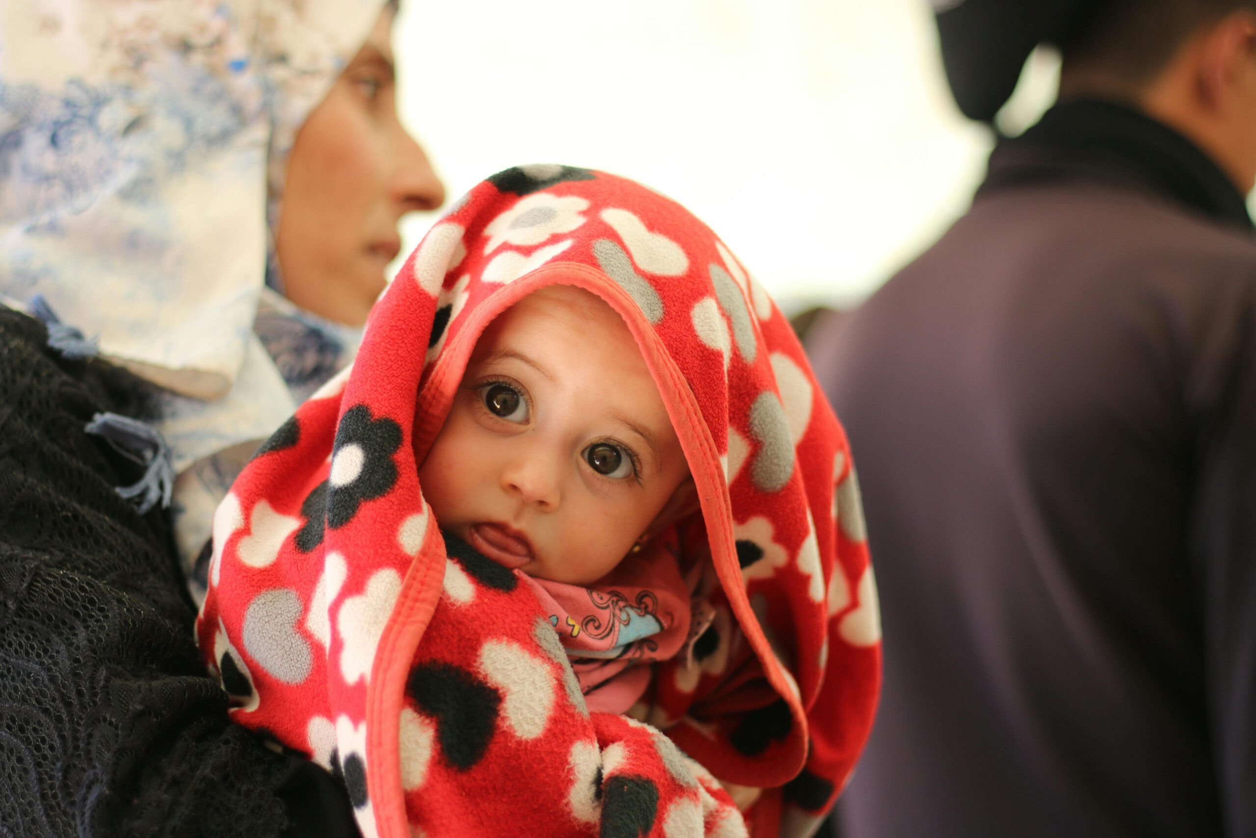 Проницательный взгляд ребенка-беженца: Жизнь за кадром миграционной статистики. Это пронзительное изображение запечатлело момент из жизни маленького человека, чья судьба неразрывно связана с миграционным кризисом. Взгляд этого ребенка, устремленный прямо в объектив, говорит громче любых цифр и отчетов. Закутанный в яркое красное одеяло с узорами, он кажется защищенным, но в то же время невероятно уязвимым. Этот снимок – напоминание о том, что за сухой статистикой «числа прошений об убежище» стоят реальные люди, их истории, страхи и надежды на мирное будущее. В контексте статьи о рекордно низком количестве заявок, это фото заставляет задуматься о цене принятых решений и о том, как меняется миграционная политика на самом деле. Это не просто иллюстрация, а визуальный комментарий к сложной и многогранной теме беженцев в Германии.