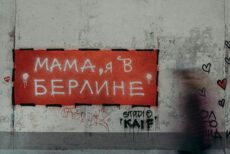 Крупный план участка шероховатой, выветрившейся бетонной стены с граффити. На стене висит прямоугольный красный знак с надписью на русском языке «МАМА, Я В БЕРЛИНЕ», выполненной белыми буквами. По краям знака — черная штриховая рамка и потеки белой краски. В правой части изображения — размытый силуэт человека в движении. На стене справа от знака — черные контуры сердец и надписи, внизу слева — небольшой стикер в виде красного сердца. Крупный план участка шероховатой, выветрившейся бетонной стены с граффити. На стене висит прямоугольный красный знак с надписью на русском языке «МАМА, Я В БЕРЛИНЕ», выполненной белыми буквами. По краям знака — черная штриховая рамка и потеки белой краски. В правой части изображения — размытый силуэт человека в движении. На стене справа от знака — черные контуры сердец и надписи, внизу слева — небольшой стикер в виде красного сердца.