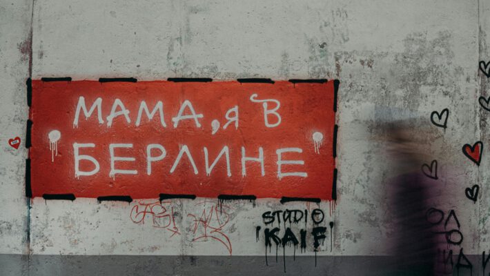 Крупный план участка шероховатой, выветрившейся бетонной стены с граффити. На стене висит прямоугольный красный знак с надписью на русском языке «МАМА, Я В БЕРЛИНЕ», выполненной белыми буквами. По краям знака — черная штриховая рамка и потеки белой краски. В правой части изображения — размытый силуэт человека в движении. На стене справа от знака — черные контуры сердец и надписи, внизу слева — небольшой стикер в виде красного сердца.
