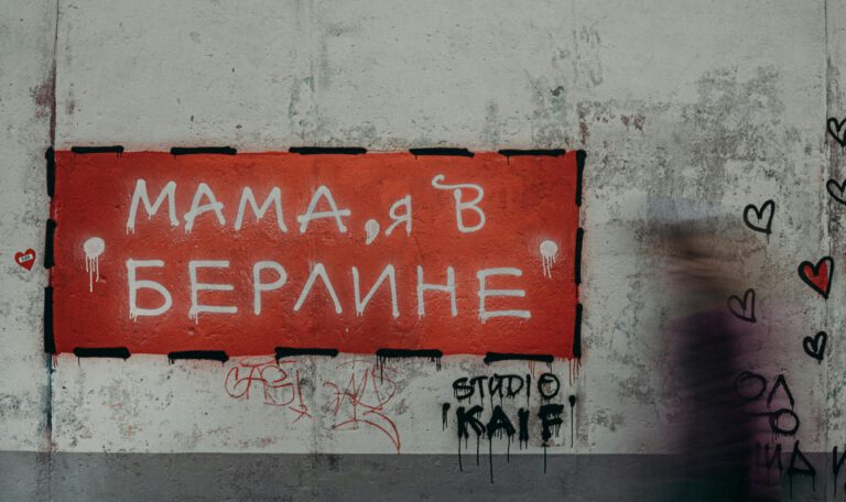 Крупный план участка шероховатой, выветрившейся бетонной стены с граффити. На стене висит прямоугольный красный знак с надписью на русском языке «МАМА, Я В БЕРЛИНЕ», выполненной белыми буквами. По краям знака — черная штриховая рамка и потеки белой краски. В правой части изображения — размытый силуэт человека в движении. На стене справа от знака — черные контуры сердец и надписи, внизу слева — небольшой стикер в виде красного сердца.