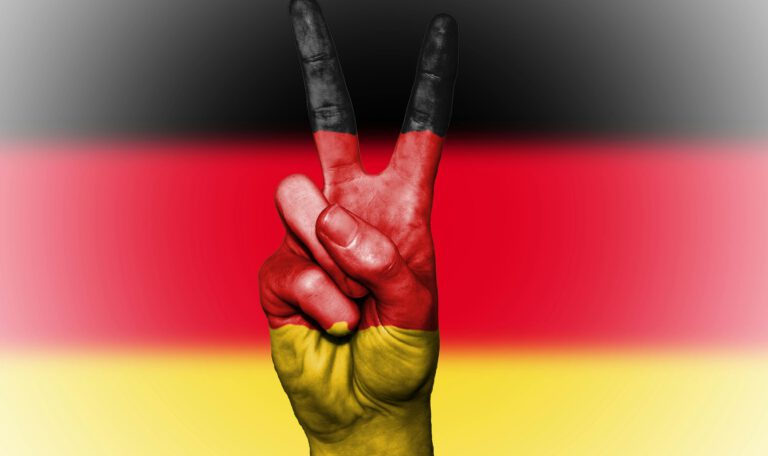 Крупный план руки, поднятой в жесте «Победа» (Peace/Victory), полностью раскрашенной в цвета флага Германии (черный, красный, золотой), на фоне размытого немецкого флага. Это изображение визуализирует дилемму «защита против мира», обсуждаемую в статье о кризисе рекрутинга в Бундесвере, и символизирует мирный, но гордый настрой немецкого общества в контексте дебатов о военной службе и патриотизме в 2026 году