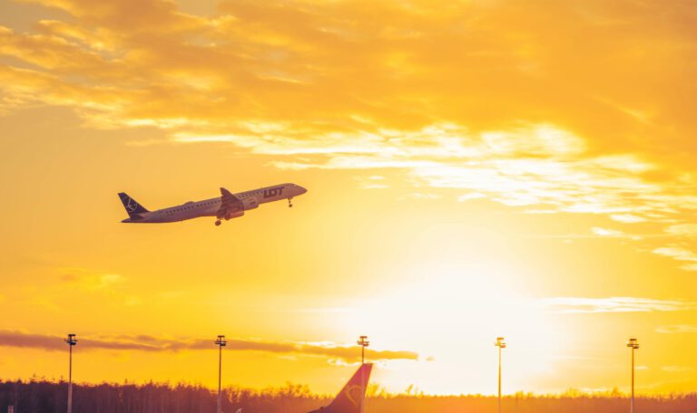 Самолет LOT Polish Airlines взлетает над аэропортом на фоне заката. Рядом с ним на взлетной полосе находится самолет Ryanair. На небе много желтых и оранжевых облаков