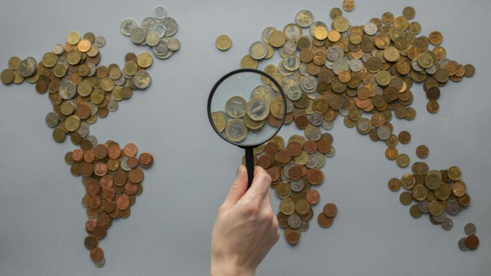 Крупный план: Рука держит черную лупу, наводя её на карту мира, сложенную из сотен разнообразных монет на сером фоне. Под увеличением лупы видны крупные монеты евро (1 и 2 евро), что символизирует детальный анализ европейской экономики и денежного обращения. Вокруг — россыпь мелких монет различных номиналов, образующих очертания континентов, демонстрируя глобальный масштаб темы и фокус на мелких деньгах.