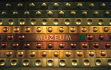 Крупный план металлической стены с надписью «MUZEUM» в современном стиле. Стену украшают ряды золотистых и медных полусферических выступов, создающих индустриальный и строгий вид, характерный для военно-технических объектов или современных музейных комплексов