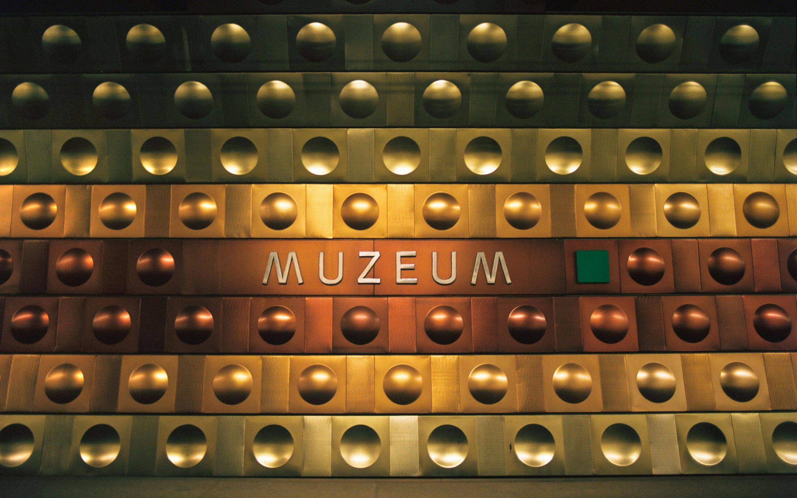 Крупный план металлической стены с надписью «MUZEUM» в современном стиле. Стену украшают ряды золотистых и медных полусферических выступов, создающих индустриальный и строгий вид, характерный для военно-технических объектов или современных музейных комплексов