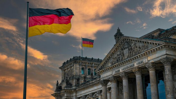 Архитектурный вид на здание Рейхстага в Берлине с надписью 'DEM DEUTSCHEN VOLKE' и колоннами. На переднем плане на высоком флагштоке развевается немецкий флаг (черно-красно-золотой), на заднем плане виден второй флаг. Небо окрашено в теплые оранжевые и розовые тона заката. Эта фотография иллюстрирует новость о правилах выезда для немцев