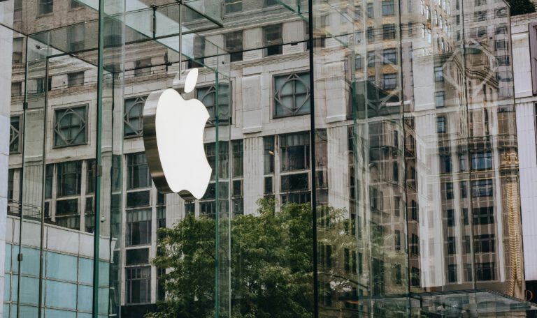 Логотип Apple на стеклянном фасаде современного здания Apple Store, в котором отражается городская архитектура и деревья. Тим Кук уходит с поста гендиректора Apple.