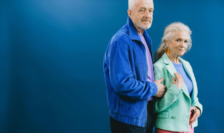 Стильный студийный портрет уверенной пожилой пары. Мужчина с седыми волосами и аккуратной бородой, одетый в ярко-синюю вельветовую куртку поверх сиреневого топа, нежно держит за руку женщину. Женщина, с длинными седыми волосами и крупными геометрическими желтыми серьгами, одета в текстурированный мятно-зеленый пиджак поверх сиреневого трикотажного свитера и розовые твидовые брюки. Сцепленные руки и прямой, спокойный взгляд на камеру представлены на глубоком, однородном синем фоне. Фото олицетворяет пенсионную систему Германии