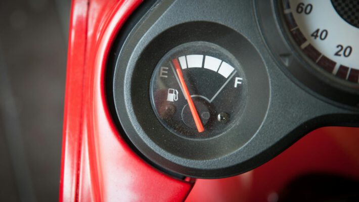 Крупный план датчика уровня топлива на приборной панели скутера или мотоцикла. Красная стрелка показывает, что бензина почти нет — она находится рядом с отметкой «E» (Empty — Пусто). Виден красный пластиковый корпус.
