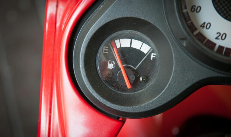 Крупный план датчика уровня топлива на приборной панели скутера или мотоцикла. Красная стрелка показывает, что бензина почти нет — она находится рядом с отметкой «E» (Empty — Пусто). Виден красный пластиковый корпус.