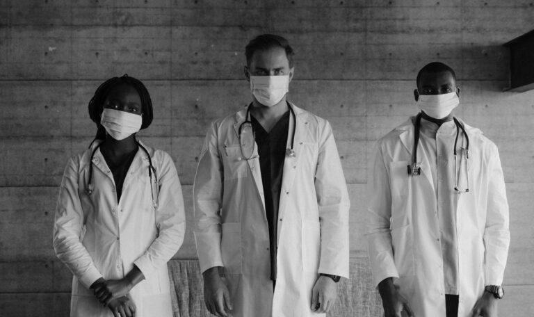 Группа из трех врачей разных национальностей в защитных масках и белых халатах в больнице перед серой бетонной стеной. Сокращение выплат по больничным