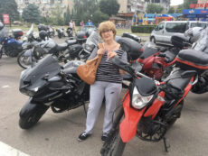 Достопримечательности Дрездена за неделю. Женщина со светлыми волосами (стрижка каре с челкой) и в очках в темной оправе стоит лицом к камере, слегка улыбаясь. Она одета в повседневную одежду: трикотажную кофту в горизонтальную черно-белую полоску, серые спортивные штаны свободного кроя и темные кеды на белой подошве. На плече у нее висит большая кожаная сумка горчично-коричневого цвета, которую она придерживает руками на уровне груди. Женщина стоит прямо между двумя мотоциклами на переднем плане. Слева от нее (для зрителя) расположен массивный спортивный мотоцикл черного цвета с надписью «Kawasaki Ninja». Справа от нее — ярко-красный мотоцикл класса эндуро или кросс. На заднем плане и по бокам видна большая парковка, полностью заполненная другими мотоциклами разных моделей и цветов (серые, синие, красные), у многих из которых сзади установлены кофры для багажа.