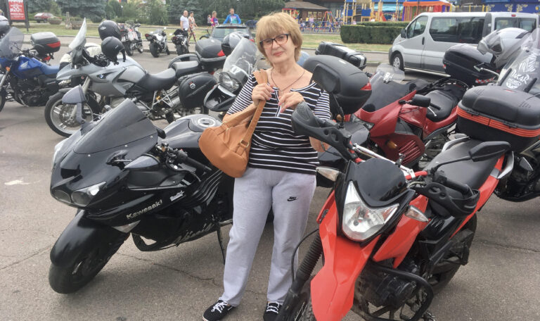 Достопримечательности Дрездена за неделю. Женщина со светлыми волосами (стрижка каре с челкой) и в очках в темной оправе стоит лицом к камере, слегка улыбаясь. Она одета в повседневную одежду: трикотажную кофту в горизонтальную черно-белую полоску, серые спортивные штаны свободного кроя и темные кеды на белой подошве. На плече у нее висит большая кожаная сумка горчично-коричневого цвета, которую она придерживает руками на уровне груди. Женщина стоит прямо между двумя мотоциклами на переднем плане. Слева от нее (для зрителя) расположен массивный спортивный мотоцикл черного цвета с надписью «Kawasaki Ninja». Справа от нее — ярко-красный мотоцикл класса эндуро или кросс. На заднем плане и по бокам видна большая парковка, полностью заполненная другими мотоциклами разных моделей и цветов (серые, синие, красные), у многих из которых сзади установлены кофры для багажа.