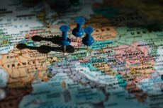 Крупний план політичної карти Європи, на якій синіми канцелярськими кнопками позначено кілька локацій. Кнопки встромлені в центральній частині карти: в районі Німеччини (біля Мюнхена), Австрії (неподалік Відня) та на кордоні Австрії з Угорщиною. На карті чітко видно назви європейських країн та міст, таких як Франція, Німеччина, Австрія, Угорщина, Румунія, а також столиці — Париж, Лондон, Брюссель, Відень, Будапешт і Бухарест. Освітлення м'яке, сфокусоване на центральній групі кнопок, через що від них падають довгі чіткі тіні в лівий бік. Навколишні краї карти злегка розмиті