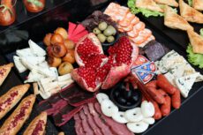 Продуктовый туризм. Разнообразная мясная и сырная закусочная тарелка (фуд-бокс) крупным планом. В самом центре композиции расположен крупный спелый гранат, разломанный на части, с ярко-красными блестящими зернами. Вокруг него аккуратными группами разложены различные закуски: ломтики белого сыра с зеленью, треугольники плавленого сыра в фольге, чечил (сыр-косичка) и круглые белые кусочки сыра. Мясная нарезка представлена вяленым мясом (бастурма), тонкими слайсами ветчины, сервелата и маленькими охотничьими колбасками. Также на тарелке присутствуют маленькие круглые плоды (вероятно, мушмула или маринованные груши), пиалы с зелеными и черными оливками. По краям видны элементы выпечки: слоеные треугольные пирожки на листьях салата и продолговатые булочки, посыпанные специями. Блюдо выглядит празднично и аппетитно, преобладают контрастные красные, белые и коричневые тона.