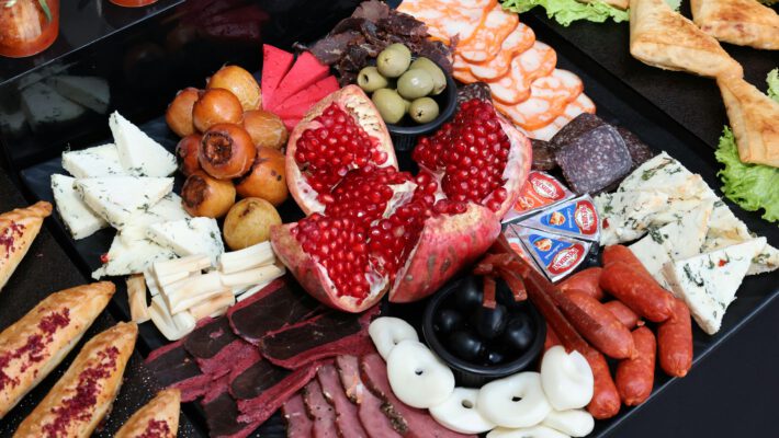 Продуктовый туризм. Разнообразная мясная и сырная закусочная тарелка (фуд-бокс) крупным планом. В самом центре композиции расположен крупный спелый гранат, разломанный на части, с ярко-красными блестящими зернами. Вокруг него аккуратными группами разложены различные закуски: ломтики белого сыра с зеленью, треугольники плавленого сыра в фольге, чечил (сыр-косичка) и круглые белые кусочки сыра. Мясная нарезка представлена вяленым мясом (бастурма), тонкими слайсами ветчины, сервелата и маленькими охотничьими колбасками. Также на тарелке присутствуют маленькие круглые плоды (вероятно, мушмула или маринованные груши), пиалы с зелеными и черными оливками. По краям видны элементы выпечки: слоеные треугольные пирожки на листьях салата и продолговатые булочки, посыпанные специями. Блюдо выглядит празднично и аппетитно, преобладают контрастные красные, белые и коричневые тона.