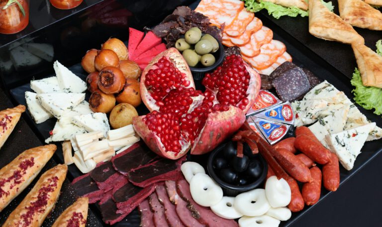 Продуктовый туризм. Разнообразная мясная и сырная закусочная тарелка (фуд-бокс) крупным планом. В самом центре композиции расположен крупный спелый гранат, разломанный на части, с ярко-красными блестящими зернами. Вокруг него аккуратными группами разложены различные закуски: ломтики белого сыра с зеленью, треугольники плавленого сыра в фольге, чечил (сыр-косичка) и круглые белые кусочки сыра. Мясная нарезка представлена вяленым мясом (бастурма), тонкими слайсами ветчины, сервелата и маленькими охотничьими колбасками. Также на тарелке присутствуют маленькие круглые плоды (вероятно, мушмула или маринованные груши), пиалы с зелеными и черными оливками. По краям видны элементы выпечки: слоеные треугольные пирожки на листьях салата и продолговатые булочки, посыпанные специями. Блюдо выглядит празднично и аппетитно, преобладают контрастные красные, белые и коричневые тона.