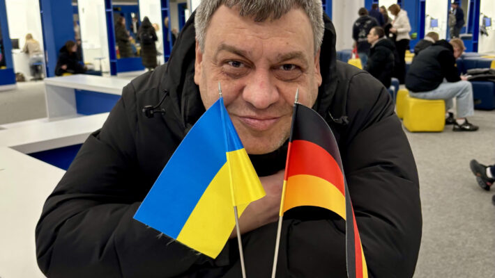 Великий план чоловіка в чорній куртці, який доброзичливо дивиться в камеру, спираючись на світлий стіл. Перед ним встановлено два маленькі настільні прапорці — прапор України та прапор Німеччини. Дія відбувається в просторому сучасному приміщенні, схожому на центр обслуговування або залу очікування, із синіми колонами та людьми на задньому плані