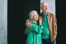 На фотографії зображена ніжна та стильна літня пара, яка стоїть, обійнявшись. Чоловік із сивим волоссям та акуратною бородою дивиться вбік, посміхаючись. На ньому яскраве зелене поло, коричнева шкіряна куртка та сині штани. Його рука лежить на плечі жінки. Жінка з довгим сивим волоссям, зібраним ззаду, ніжно дивиться на чоловіка. На ній довга сукня насиченого зеленого кольору з об’ємними рукавами та довгі сережки. Її руки лежать поверх руки чоловіка. Вони стоять на тлі стіни, пофарбованої в чорний колір, яка обрамлена сірими колонами з грубою текстурою. Освітлення м’яке і тепле. Фотографія передає атмосферу любові, поваги та довгих стосунків, що є особливо актуальним для майбутніх пенсіонерів, які мають значний **трудовий стаж до 2004 року** та готуються до заслуженого відпочинку.